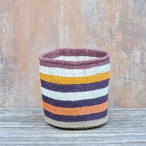 May include: A cylindrical woven basket with horizontal stripes in shades of purple, orange, yellow, and white. The basket has a natural, neutral-coloured base and a darker purple rim. The basket is made of natural fibres.