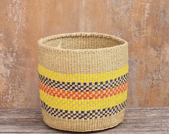 May include: A cylindrical woven basket made of natural tan fibers, featuring horizontal bands of bright yellow, dark charcoal, and vibrant orange woven patterns around the center, with a wide open top and a flat, circular base structure.