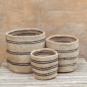 May include: Three woven baskets of varying sizes. The baskets are cylindrical with a natural tan color and horizontal black stripes. They are arranged on a wooden surface against a neutral background.
