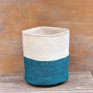 May include: A cylindrical woven basket featuring a two-tone design with an off-white upper section and a deep teal lower section. The basket has a consistent, tight weave pattern throughout its structure, creating a sturdy, upright, open-top container.