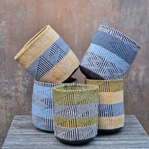 Può includere: Una collezione di cesti portaoggetti in tessuto intrecciato in vari colori, tra cui giallo, blu e grigio. I cesti presentano motivi geometrici e sono disposti su una superficie di legno. I cesti sono cilindrici.