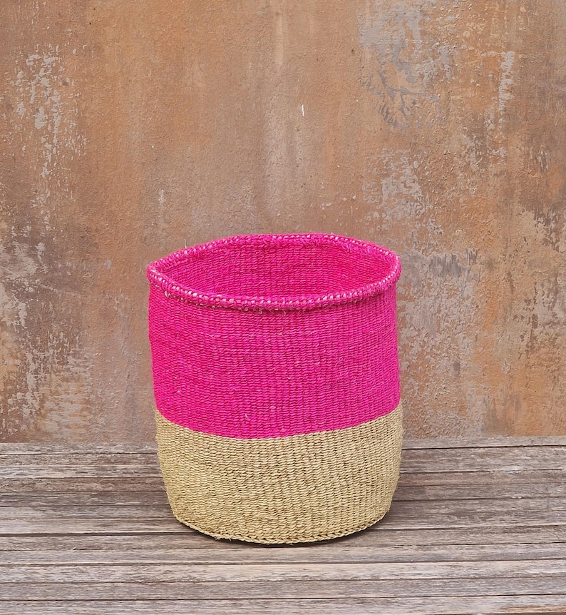 May include: A handwoven basket with a cylindrical shape. The top half is a vibrant pink, while the bottom half is a natural tan color. The basket is made of woven fibers and sits on a wooden surface.