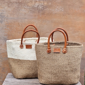 May include: Two woven tote bags with leather handles. The bag on the left is white with a brown bottom. The bag on the right is all brown. Both bags have a leather tag that says "Kenyan Craft".