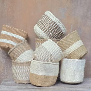 May include: A collection of woven baskets in various sizes, featuring natural tan and off-white colour palettes. The baskets display diverse patterns including horizontal stripes, solid blocks of colour, and intricate textured weave designs.
