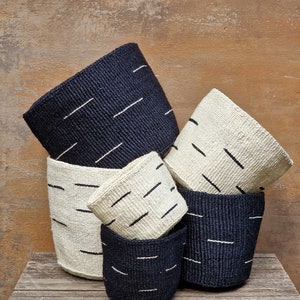 May include: A set of four woven baskets in black and white. The baskets are stacked on top of each other, with the largest basket at the bottom and the smallest basket at the top. The baskets are made from a natural material and have a simple, geometric design.