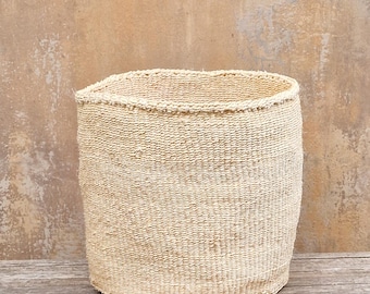 May include: A cylindrical, beige woven basket with a slightly irregular top edge. The basket is made of a natural fiber and has a textured surface. It sits on a wooden surface against a neutral-toned wall.