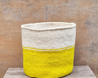 May include: A woven basket with a cylindrical shape. The top half is a natural off-white color, while the bottom half is bright yellow. A thin yellow stripe separates the two colors. The basket sits on a wooden surface.