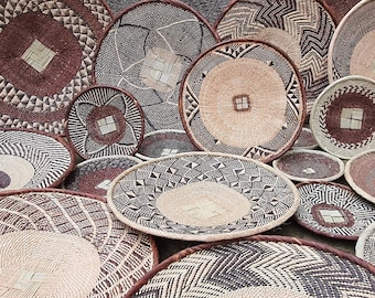 May include: A collection of round, woven wall baskets featuring intricate geometric patterns in shades of brown, black, and natural tan. Each basket displays a distinct design with a central square weave detail and a reinforced, dark brown rim.