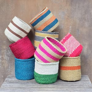 May include: A collection of woven baskets in various colors and patterns, including solid blue, solid pink, green and white, tan with orange stripes, and multi-colored striped designs. The baskets are stacked together in a group, showcasing different textures.