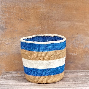 May include: A cylindrical woven basket featuring horizontal stripes in vibrant blue, natural tan, and off-white. The basket has a sturdy, open-top design with a consistent, textured weave pattern throughout the exterior and interior surfaces.