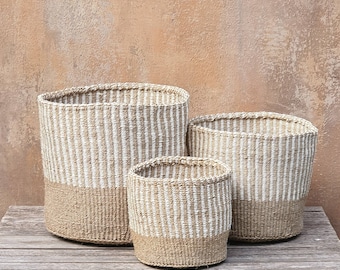 Puede incluir: Tres cestos de almacenamiento tejidos de diferentes tamaños. Los cestos son de color beige con rayas blancas verticales en la parte superior y una base beige sólida. Están colocados sobre una superficie de madera contra un fondo texturizado de tonos neutros.