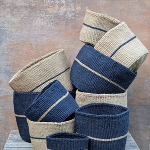 Puede incluir: Un conjunto de cestas tejidas de varios tamaños, de color beige natural y rayas azules. Las cestas están apiladas y dispuestas en forma de pirámide.