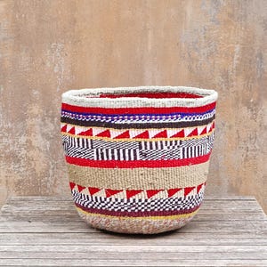 Könnte beinhalten: Ein geflochtener Korb mit horizontalen Farbbändern in Rot, Weiß, Schwarz und Naturbeige. Das Design umfasst geometrische Muster mit sich wiederholenden Dreiecken und karierten Streifen auf der gesamten Außenfläche des runden Behälters.