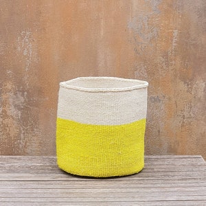 May include: A cylindrical woven basket with a flat base. The top half is a natural off-white color, while the bottom half is a bright yellow. The basket is sitting on a wooden surface against a textured brown background.