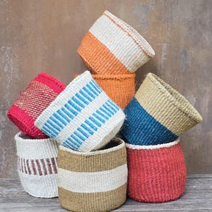 May include: A collection of eight cylindrical woven baskets in various colors including red, orange, blue, white, and tan. The baskets feature diverse patterns such as solid blocks, horizontal stripes, and vertical dashes, all crafted with a textured fiber.