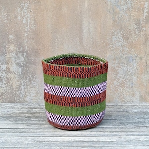 May include: A cylindrical woven basket featuring horizontal stripes in olive green, dark brown, and light pink. The basket has a textured, multi-colored weave pattern throughout its exterior, creating a distinct banded appearance from top to bottom.