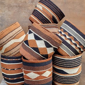 May include: A collection of cylindrical woven baskets featuring geometric patterns in black, white, and terracotta orange. The baskets are stacked together, displaying various horizontal stripes, diamond shapes, and textured weave designs.
