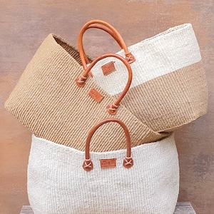 Op de afbeelding: Drie geweven manden met leren handvatten. De manden zijn op elkaar gestapeld. De bovenste mand is wit, de middelste mand is bruin en de onderste mand is wit. Elke mand heeft een leren label met het woord "Kenyan" erop.