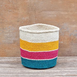 May include: A woven basket with a cylindrical shape and a slightly oval top. The basket features horizontal stripes in cream, yellow, fuchsia, and teal. The basket is made of natural fibers and sits on a wooden surface.