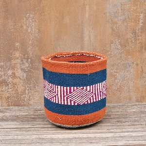 May include: A cylindrical woven basket featuring horizontal bands of burnt orange and deep blue. A central section displays a geometric pattern in magenta and cream. The basket has a thick, textured weave and an open top with a visible rim.