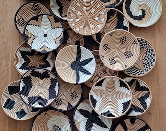 May include: A collection of round, woven wall baskets featuring intricate geometric patterns in black, white, and natural tan. Each basket displays a unique design, including star shapes, spirals, and checkered motifs, crafted from coiled fibers.