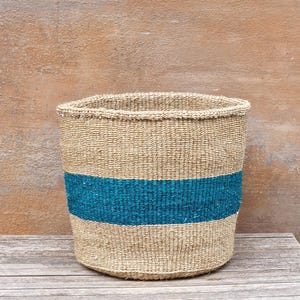 May include: A cylindrical woven basket in natural beige, featuring a broad teal blue stripe. The basket has a slightly flared top and is crafted from a textured, natural fibre.