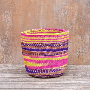 May include: A woven basket with a cylindrical shape. The basket features horizontal stripes in vibrant colors, including pink, yellow, purple, and brown. The basket is made of natural fibers and sits on a wooden surface.