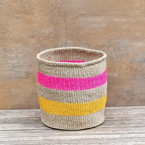 May include: A cylindrical woven basket with a natural beige base, accented by horizontal bands of bright pink and yellow. The basket is placed on a wooden surface against a neutral background.