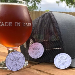 May include: A gray and black mesh trucker hat with a black and white circular patch that says "Some Good Hops" and a silver pin with the same logo. The hat is sitting on a wooden table next to a glass of beer and another silver pin. The glass of beer has the words "Made in Dade" etched on it.