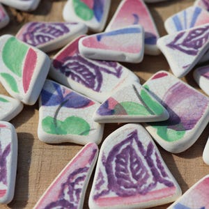 Może przedstawiać: Kolekcja nieregularnych odłamków ceramicznych z białym tłem i malowanymi wzorami w kolorach fioletowym, różowym i zielonym. Każdy element ma gładką, zaokrągloną krawędź oraz widoczną fakturę spękanej glazury na powierzchni ceramicznych fragmentów.
