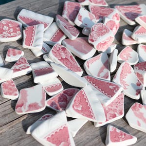 Puede incluir: Un conjunto de fragmentos de cerámica rotos, con base blanca y diseño floral rosa. Las piezas, de tamaños y formas variadas, están dispuestas sobre una superficie de madera. El conjunto tiene una estética vintage.