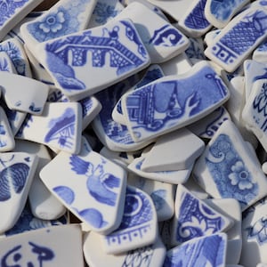 Può includere: Una collezione di frammenti di ceramica bianca con vari motivi di trasferimento blu, tra cui raffigurazioni di ponti, motivi floreali e uccelli. Ogni pezzo ha una forma irregolare con bordi lisci, mostrando un mix di disegni complessi.