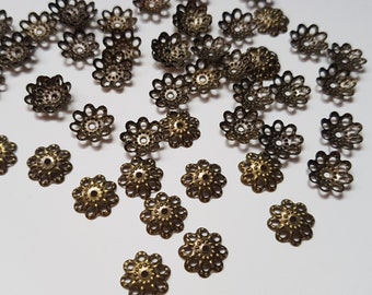 May include: A collection of gold-toned metal bead caps with a decorative flower design. The caps are scattered on a white surface.