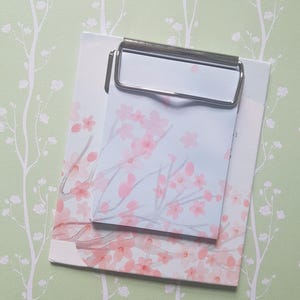 Könnte beinhalten: Ein rechteckiger Notizblock mit einer zarten Aquarellillustration von rosa Kirschblüten auf weißem Papier. Der Notizblock ist oben mit einer polierten silbernen Metallklammer befestigt, die ein einfaches Entfernen der einzelnen Seiten ermöglicht.