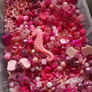 Può includere: Un vasto assortimento di perline rosa, ciondoli e altri materiali per l'artigianato in un contenitore di plastica trasparente. Gli articoli includono cuori, stelle, fiori e un cavalluccio marino.