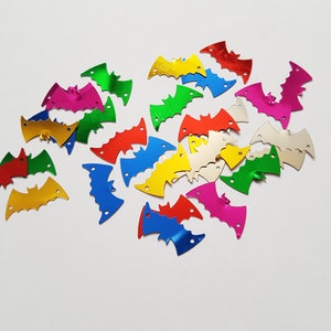 Op de afbeelding: Een verzameling kleurrijke, metalen pailletten in de vorm van vleermuizen. De pailletten zijn in verschillende kleuren, waaronder rood, oranje, geel, groen, blauw, paars en zilver.