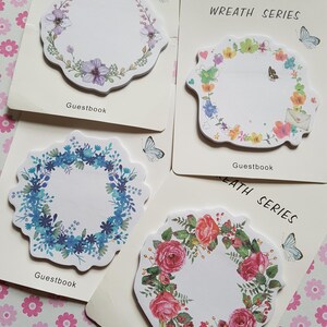 May include: Four sets of Wreath Series Guestbook sticky notes on white backing cards. Each set features a unique floral wreath design, including purple flowers, colorful wildflowers with an envelope, blue blossoms, and red roses with green leaves.