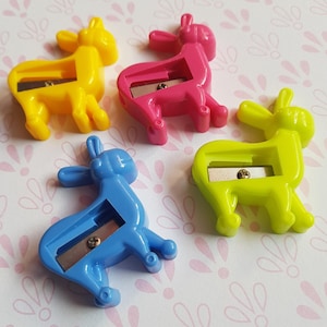 May include: Four plastic pencil sharpeners shaped like standing donkeys in bright yellow, hot pink, light blue, and neon green. Each animal-shaped sharpener features a metal blade secured by a small silver screw in the center of the body.