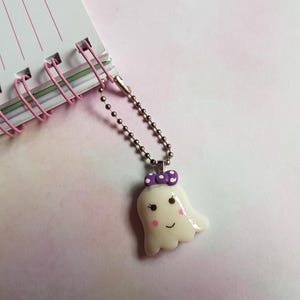Puede incluir: Un dije de fantasma blanco con un lazo morado de lunares, ojos negros y mejillas rosadas, unido a una cadena de bolas de metal plateado. El dije está conectado a la espiral rosa de un cuaderno blanco con líneas horizontales rosas finas.