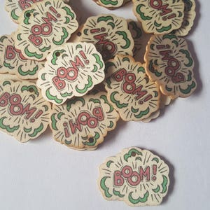 Könnte beinhalten: Ein Stapel Holz-Etiketten mit einer roten und weißen "BOOM!" Grafik auf weißem Hintergrund. Die Etiketten sind in Form einer Sprechblase mit grünen Umrissen.