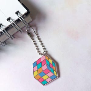 May include: A hexagonal acrylic charm featuring a colorful geometric cube pattern in shades of pink, blue, yellow, and orange. The charm is attached to a silver-toned metal ball chain, which is looped through the spiral binding of a white notebook.