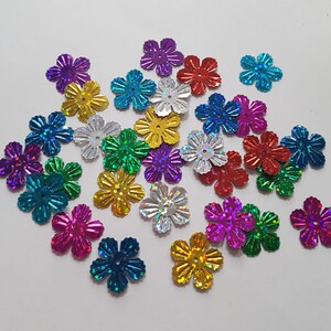 Op de afbeelding: Assortiment van kleurrijke, iriserende bloemvormige pailletten. De pailletten zijn gemaakt van een glanzend, metalen materiaal en zijn verkrijgbaar in verschillende kleuren, waaronder goud, zilver, rood, blauw, groen, paars en roze.