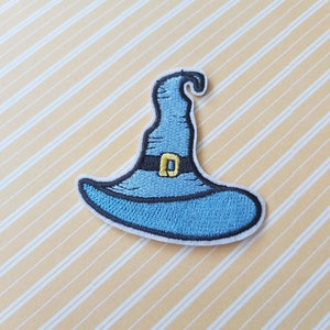 May include: A blue witch's hat with a black band and a yellow buckle. The hat is embroidered on a white fabric.
