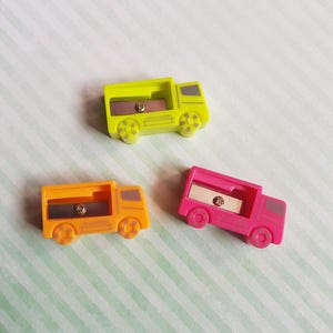 May include: Three pencil sharpeners shaped like lorries in lime green, orange, and hot pink. Each plastic sharpener features a metal blade, a screw, and a small window for pencil insertion. The word SHARPENER is embossed on the side of each lorry.