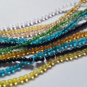 Könnte beinhalten: Sortiment an Glasperlen in verschiedenen Farben, darunter klar, gelb, grün, blau und schwarz. Die Perlen sind alle klein und haben eine leicht längliche Form.