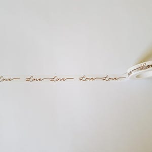 May include: A roll of white washi tape featuring a repeating pattern of the word Love written in a brown cursive script. The tape is partially unrolled, displaying the continuous text design across the length of the adhesive strip.