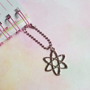 May include: A silver-toned metal charm shaped like an atom with intersecting elliptical orbits, attached to a small silver ball chain. The charm is connected to the spiral binding of a notebook with pink wire loops.