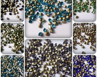 May include: Assortment of small, faceted, glass rhinestones in various colors, including black, green, blue, gold, and silver. The rhinestones are scattered on a white surface.