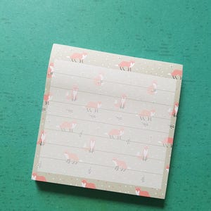 May include: A square notepad featuring a repeating pattern of small orange foxes on a light beige background. The paper is lined with thin horizontal grey lines and decorated with tiny white dots and small grey leaf icons scattered across the surface.