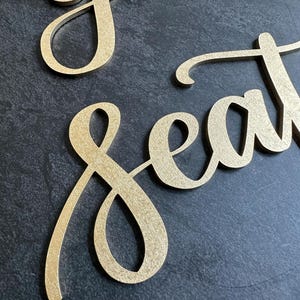 May include: Gold-colored script lettering cutouts spelling the words "your seat". The letters have a textured, metallic finish and are presented in a cursive font style with connected characters, creating a decorative sign for event seating.
